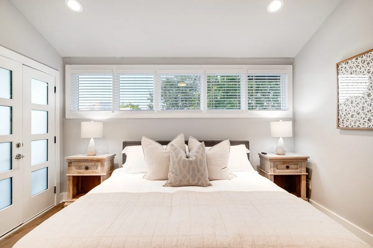 A serene bedroom featuring a bed with plush decorative pillows, nightstands with lamps on either side, and a window with blinds that allows soft natural light to filter in. The room exudes calm with its understated color palette and cozy atmosphere, ideal for relaxation and unwinding.