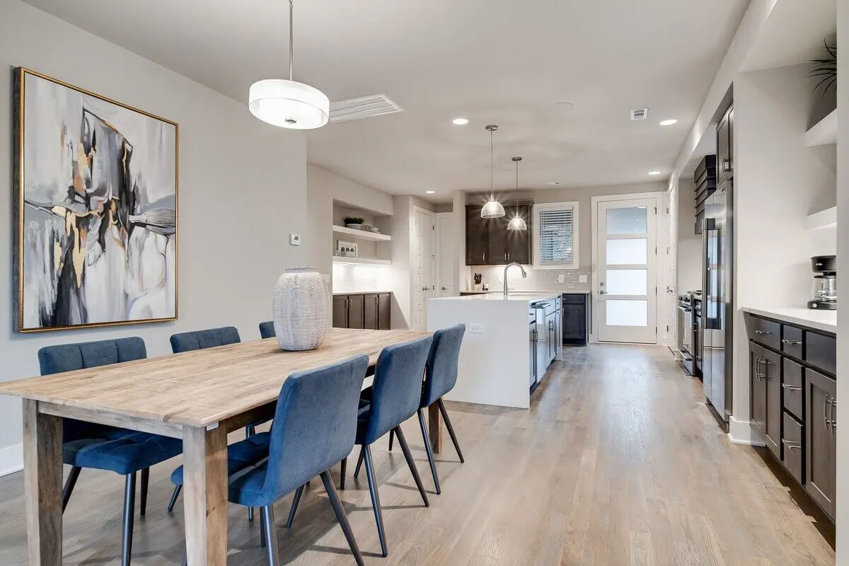 A spacious, warmly lit dining area features a large table set beneath an abstract art piece. The kitchen island is visible in the background, seamlessly connecting the dining and cooking spaces. The setting is clearly designed for both shared meals and social gatherings, with ample seating and modern décor.