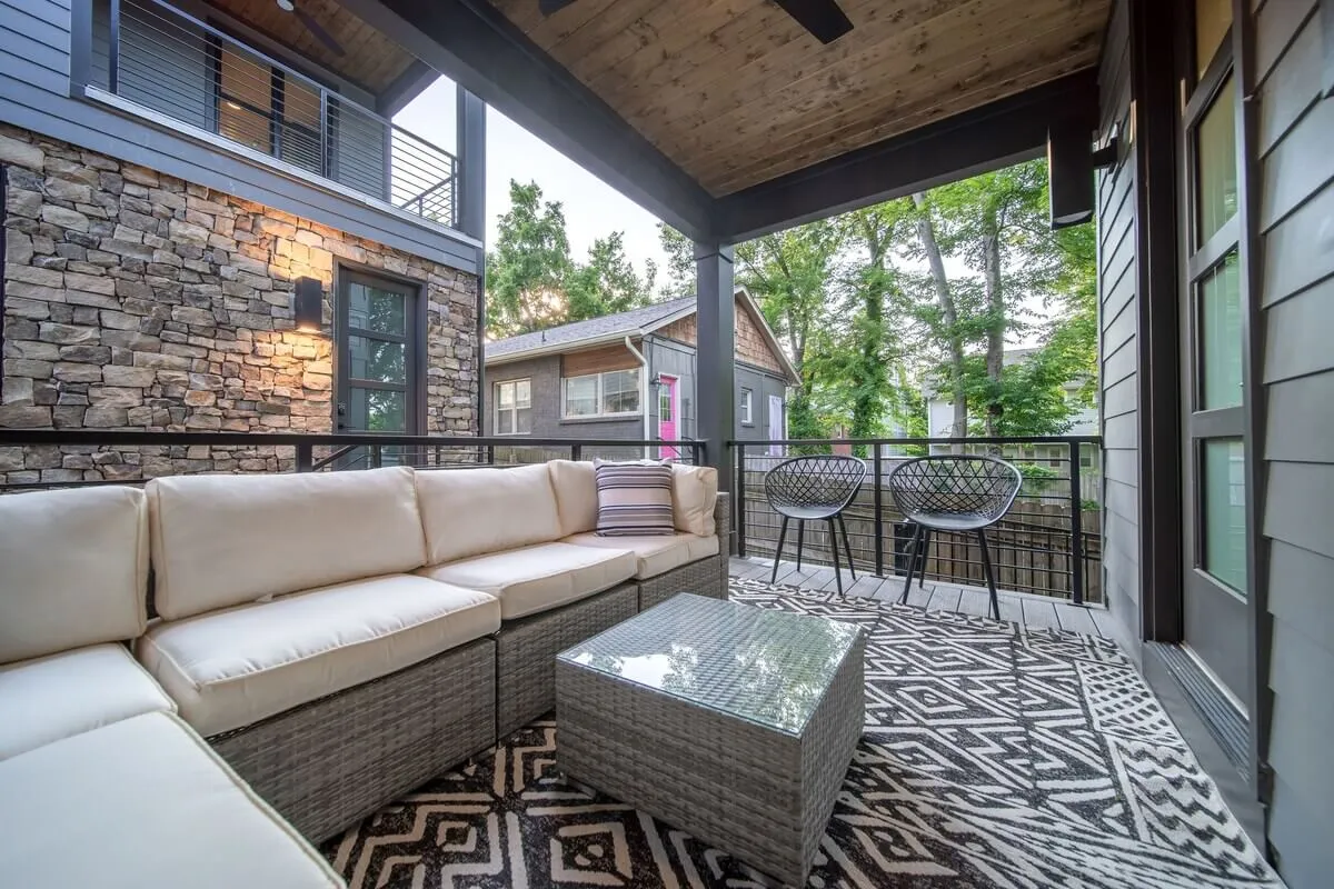 A beautifully arranged outdoor seating area featuring a glass-top coffee table, a textured rug, and comfortable chairs surrounded by lush greenery. The area is bathed in natural light and conveys a seamless transition between indoor comfort and outdoor relaxation.