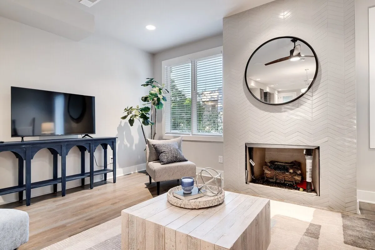 A modern living area featuring a large television set above a striking herringbone-patterned fireplace, a round mirror, and a well-appointed coffee table. The setting invites guests to relax, watch movies, or entertain friends in a sophisticated environment.