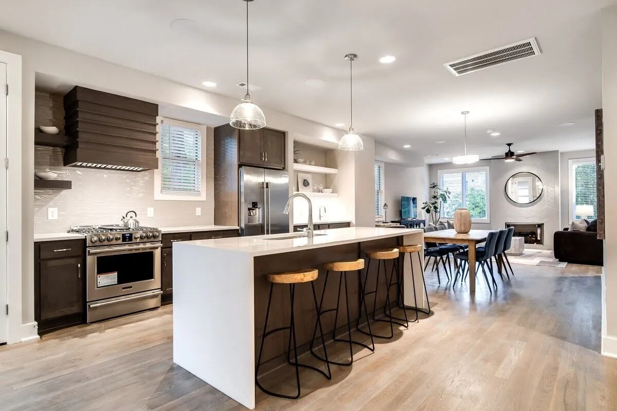 A modern kitchen with a large island accompanied by stools, opening up to a dining area and bright window views. The space is equipped with sleek appliances and offers a relaxed, contemporary vibe for both cooking and entertaining guests.