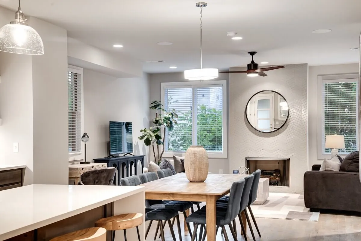 A warmly lit, open-concept interior showcasing a cozy living room with a plush sofa, a stylish decorative mirror above a fireplace, and a dining table set for gatherings. The arrangement encourages conversation and relaxation, blending chic design with comfort.