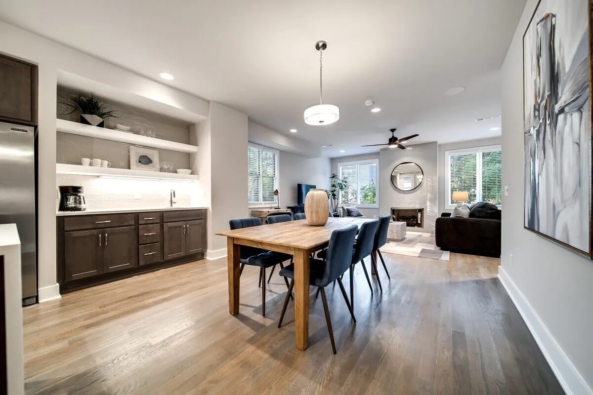 A dining area that seamlessly connects to the kitchen and living space, with large windows letting in greenery from outdoors. The scene includes a stylish dining table, comfortable couch, and decorative artwork for a warm, welcoming atmosphere.