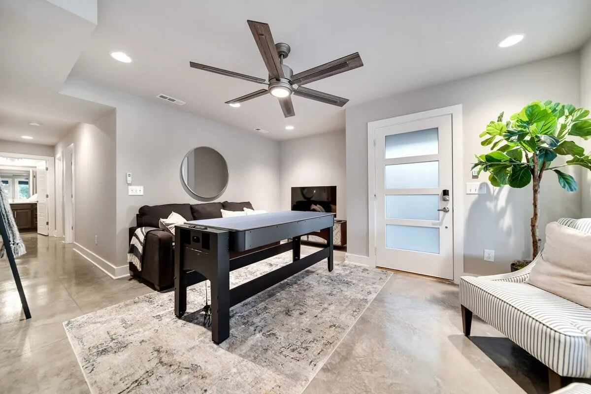 A casual, inviting living space featuring a table, comfortable sofa, lush green plant, ceiling fan, and large mirror. The setup suggests a spot for families to gather, play games, or plan the day's adventures, enhanced by natural light and cozy decor.
